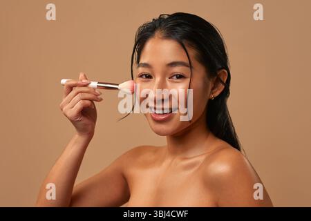 Asiatische Frau mit sauberer gesunder leuchtender Haut mit Make-up-Pinsel isoliert auf beigem Hintergrund. Schönheitskonzept, Make-up. Stockfoto