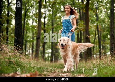 Eine fröhliche Frau in bequemen Overalls spaziert mit ihrem verspielten goldenen Retriever durch einen üppigen grünen Wald und genießt dabei die Wunder der Natur und der Natur Stockfoto