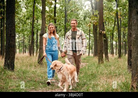 Ein fröhliches Paar schlendert Hand in Hand durch einen ruhigen Wald und genießt die Schönheit der Natur neben ihrem verspielten Golden Retriever, Experienc Stockfoto