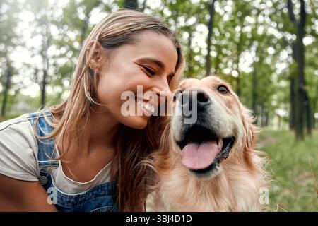 In einem ruhigen Park strahlt eine fröhliche junge Frau vor Glück, während ihr verspielter goldener Retriever neben ihr das wahre Wesen der Gemeinschaft, der Freude, verkörpert Stockfoto