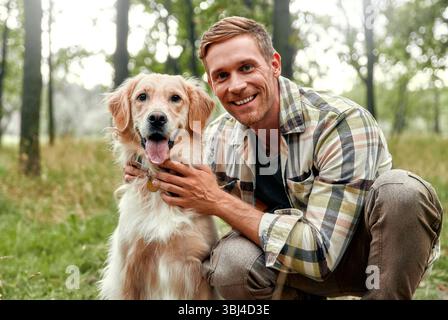 Ein fröhlicher Mann genießt einen schönen Moment mit seinem verspielten Golden Retriever in einem üppigen Wald, der Freude und Gesellschaft in der Natur einfängt, ideal für Haustiere Stockfoto