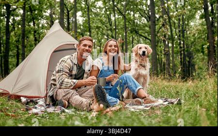 Ein fröhliches Paar genießt einen wunderbaren Campingausflug tief in den Wald mit seinem verspielten goldenen Retriever, während sie sich in der Schönheit der Natur und sonnen Stockfoto