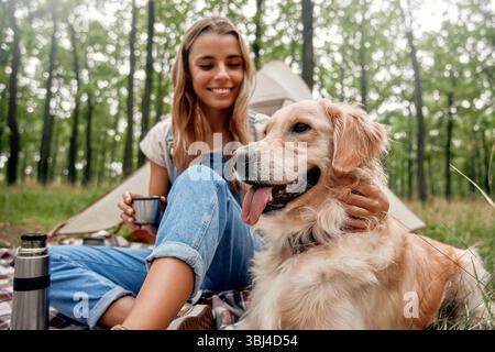 Ein fröhliches Mädchen hat einen wunderbaren Moment mit ihrem goldenen Retriever, während er einen Campingausflug in einer ruhigen und ruhigen Waldlandschaft genießt, was ich Stockfoto
