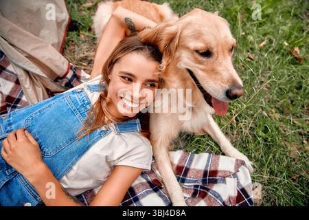 Eine junge Frau strahlt Freude und Glück aus, während sie hell lächelt, während sie sich bequem neben ihrem fröhlichen goldenen Retriever auf einer Picknickdecke ausruht, al Stockfoto