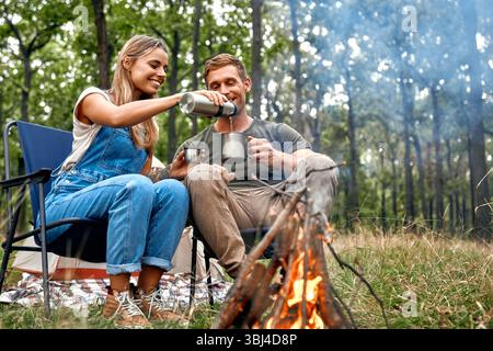 In einem üppigen, lebhaften Wald lacht ein liebevolles Paar am warmen Lagerfeuer und genießt dampfenden heißen Kaffee, während es das ruhige Breat genießt Stockfoto