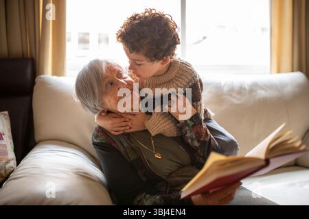 Enkel umarmt Großmutter, während sie ein Buch liest und zu Hause auf dem Sofa sitzt und die gemeinsame Familienzeit genießt Stockfoto