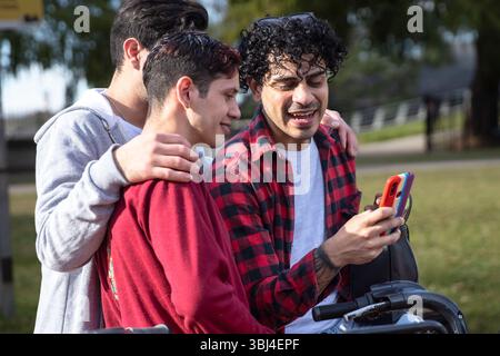 Drei Freunde lachen, während sie sich im Freien um ein Telefon sammeln und einen sonnigen Tag im Park genießen. Stockfoto