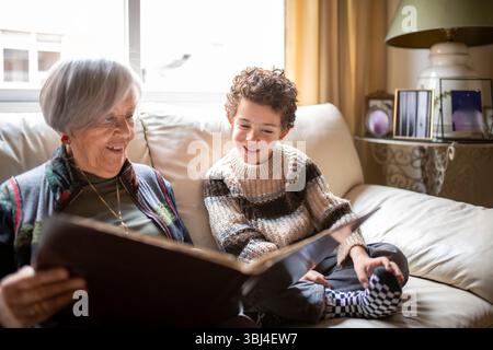 Großmutter und Enkelkind genießen eine gute gemeinsame Zeit, stöbern auf einem gemütlichen Sofa durch ein Fotoalbum und erinnern an die liebsten Erinnerungen Stockfoto