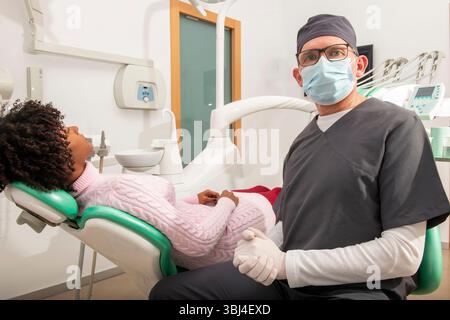 Zahnarzt mit Maske und Handschuhen posiert in seiner Klinik neben einem auf dem Zahnarztstuhl liegenden Patienten Stockfoto