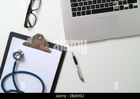 Ansicht von oben auf den Arbeitsplatz eines modernen medizinischen Fachpersonals. Das Bild zeigt einen silbernen Laptop, ein Stethoskop auf einer leeren Zwischenablage, einen Stift und Stockfoto