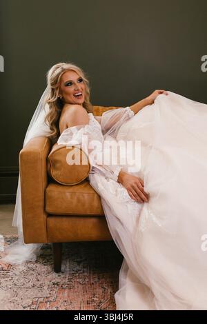 Blonde Braut mit Schleier und Hochzeitskleid auf der Couch im Wohnzimmer Stockfoto