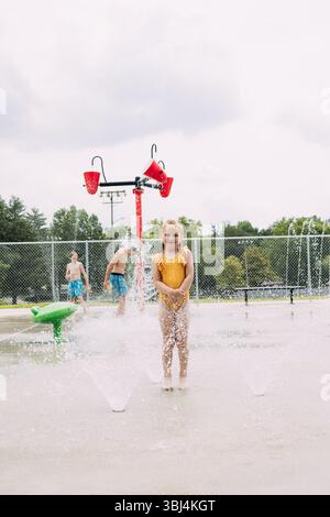 Lächelndes Kind, das in den Sprinklern steht, während die Kinder in der Planschmatte spielen Stockfoto