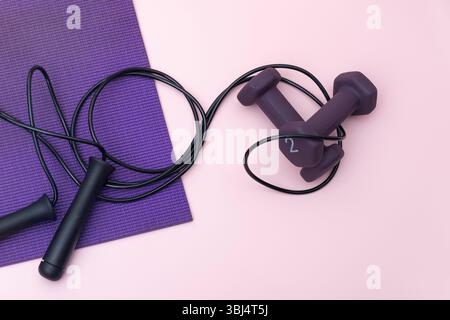 Lila Kurzhanteln, Sprungseil und Yogamatte auf rosa Hintergrund Stockfoto