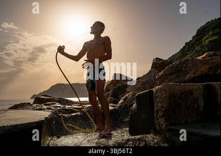 Bei Sonnenuntergang spült ein Mann mit einem Schlauch am felsigen Strand von Vernazza, Cinque Terre, Italien, unter einem dramatischen Himmel ab, der von warmem Abendlicht erstrahlt. Stockfoto