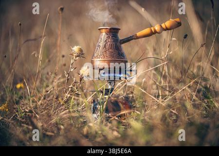Kaffee in türkischem Cezve auf Gasbrenner für Camping kochen. Feuer mit blauer Flamme und Dampf über heißem, köstlichem Kaffee zwischen Disteln. Morgenkaffee in n Stockfoto
