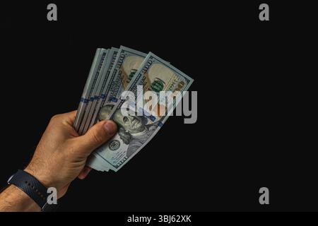 Eine Hand zeigt mehrere hundert-Dollar-Scheine vor dunklem Hintergrund, was die Struktur und Details der Währung bei schlechten Lichtverhältnissen unterstreicht. Stockfoto