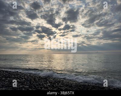 Kieselstrand mit sanften Wellen unter dramatisch bewölktem Himmel und Sonnenstrahlen über dem Schwarzen Meer in Batumi, Georgia. Natürlicher Stimmungshintergrund. Stockfoto