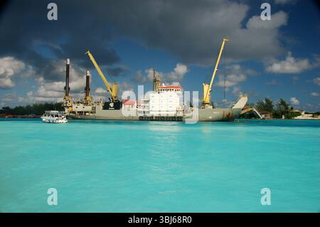 Industriegüterschiff mit großen Kränen, die in türkisfarbenem Wasser unter einem dramatischen Himmel in einem karibischen Hafen angedockt sind, mit einem kleinen Boot in der Nähe Stockfoto