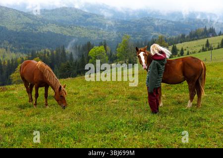 Eine ruhige Szene, in der sich ein junger Mensch auf einer wunderschönen grünen Wiese vor einer Kulisse sanfter Hügel mit zwei Pferden verbindet Stockfoto
