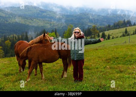 Eine Frau steht mit offenen Armen und interagiert freudig mit zwei Pferden auf einer lebhaften grünen Wiese, umgeben von majestätischen Bergen unter einer teilweise bewölkten Fläche Stockfoto