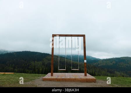 Dieses Bild zeigt eine wunderschön gefertigte Holzschaukel vor der Kulisse von sanften grünen Hügeln und einem bewölkten Himmel, die Besucher zum Genießen von Tram einlädt Stockfoto