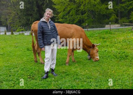 Eine junge Person in lässiger Kleidung steht neben einer grasenden braunen Kuh in einem üppigen grünen Feld, was eine ruhige Verbindung zwischen Menschen veranschaulicht Stockfoto