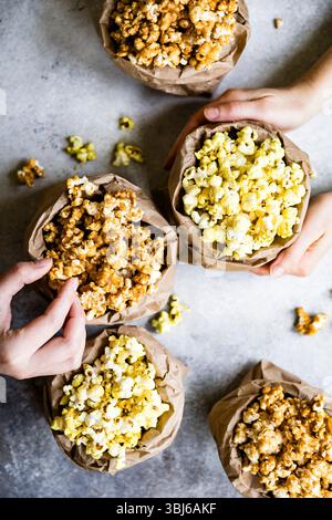 Eine Auswahl an Popcorn-Aromen in braunen Papiertüten, wobei die Hände auf einer strukturierten Oberfläche nach den Snacks greifen. Stockfoto