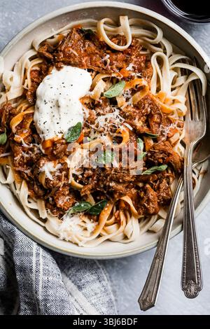 Eine Schüssel Pasta mit reichhaltiger Fleischsauce, geriebenem Käse und frischen Kräutern, begleitet von einer Gabel und Leinenserviette. Stockfoto