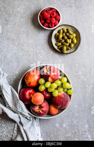 Eine Auswahl an frischen Früchten, darunter Pfirsiche, Nektarinen, Trauben und Himbeeren, zusammen mit einer Schüssel grüner Oliven auf einer strukturierten Oberfläche. Stockfoto