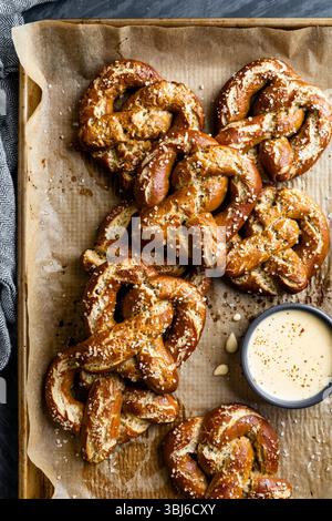 Eine Portion frisch gebackener Brezeln mit grobem Salz wird auf einem mit Pergament gefütterten Tablett präsentiert, begleitet von einer kleinen Schüssel cremiger Dip-Sauce. Stockfoto