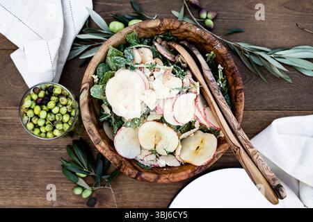 Eine hölzerne Schüssel gefüllt mit einem frischen Salat mit geschnittenen Äpfeln, Gemüse und Nüssen, begleitet von einem Glas gemischter Oliven auf einem Holztisch. Stockfoto