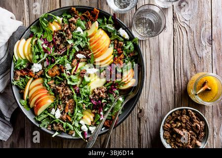 Ein frischer Salat mit geschnittenen Äpfeln, Rucola, Radicchio, Pekannüssen, Ziegenkäse, und ein Dressing wird in einer dunklen Schüssel auf einem Holztisch serviert. Stockfoto