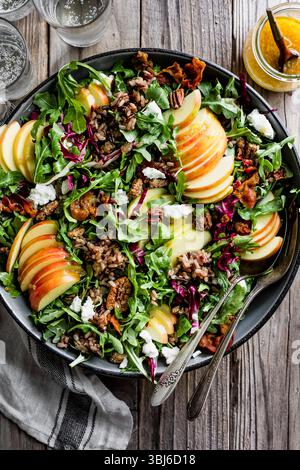 Ein lebendiger Salat mit Rucola, Apfelscheiben, Ziegenkäse, Pekannüssen und gemischten Körnern, serviert in einer dunklen Schüssel auf einem Holztisch. Stockfoto