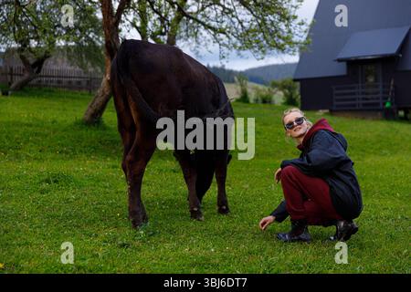 Eine fröhliche junge Frau, die eine Sonnenbrille und ein gemütliches Outfit trägt, genießt einen ruhigen Moment mit einer Kuh in einem üppigen grünen Feld, umgeben von Bäumen und einem stilvollen Mod Stockfoto