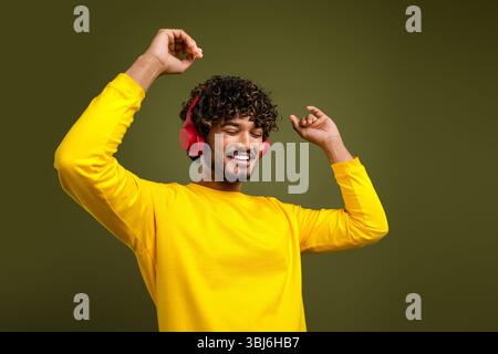 Ein fröhlicher junger Mann, der Musik mit kabellosen Kopfhörern genießt und trendige gelbe Kleidung auf einem grün leuchtenden Hintergrund trägt Stockfoto
