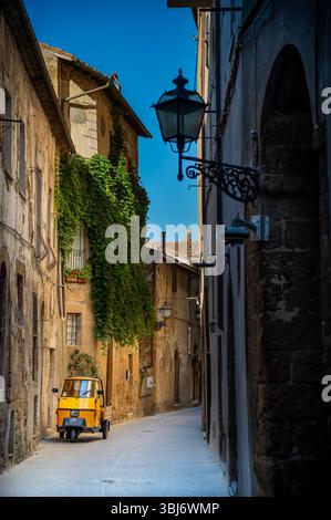 Ein gelbes dreirädriges Ape-Fahrzeug, das auf einer leeren Kopfsteinpflasterstraße in der mittelalterlichen Stadt Pitigliano in der Toskana, Italien, geparkt ist – bezaubernder und vertikaler Blick Stockfoto