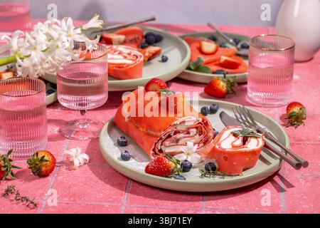 Köstliche rote Erdbeeren gerollte Pfannkuchen serviert auf leuchtend rosa Tellern mit frischen Erdbeeren, Heidelbeeren und zarten weißen Blüten, gepaart mit r Stockfoto