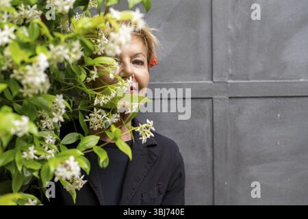 Kaukasische ältere elegante, elegante, wohlhabende Frau mit kurzen blonden Haaren und schwarzer Jacke, die sich neben einer grauen Wand versteckt, während sie sich hinter einem blühenden Jasmin versteckt Stockfoto