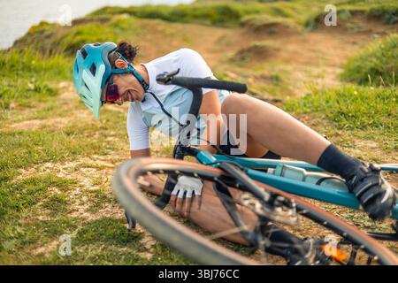 Lateinische Radfahrerin fällt von ihrem Schotterrad und berührt ihr Knie nach einem Unfall während des Trainings auf einem unbefestigten Weg am Meer bei Sonnenuntergang Stockfoto
