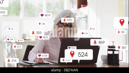 Beratung einer Seniorin, die Laptop scannt und in Notizblock am Küchentisch schreibt, mit Benachrichtigungen Stockfoto