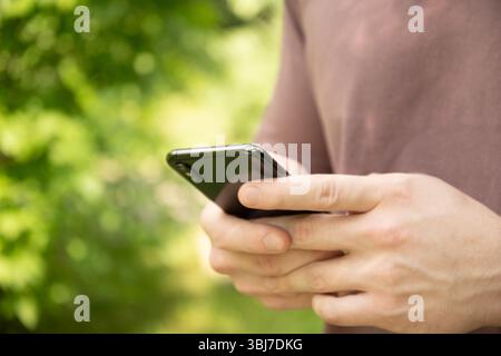Nahaufnahme der Hände des Mannes, die das Smartphone im grünen Außenbereich halten. Schauen Sie sich Apps an, die sich auf Geräte, Chats, SMS oder Internet-Browsen konzentrieren. Stockfoto