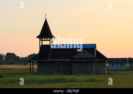 Alte Holzkirche mit hohem Turm und Kreuz steht auf einem offenen Feld, Dach teilweise mit blauer Plane bedeckt, verwitterte Bretter und geschlossene Rollläden Stockfoto