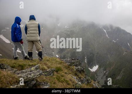 Zwei Wanderer mit Trekkingstöcken stehen auf nebeligem Bergrücken und betrachten das dramatische Alpental mit Schneestücken und nebeligem Wetter. Stockfoto