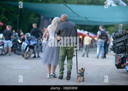 Ein Paar spaziert mit ihrem kleinen Hund beim Moto Rock fest 2025 in Ruse, Bulgarien. Der Mann trägt eine Lederjacke und Camouflage-Hose und lehnt sich an seinen Partner, der in einem hellgrauen Tüllrock und weißen Sneakers gekleidet ist, mit einem Gehstock. Ein Yorkshire Terrier trabt neben ihnen. Um sie herum erkunden die Besucher den Ausstellungsbereich für Motorräder, während verschiedene Fahrräder unter großen Sonnendächern parken. Die Abendszene verbindet Zuneigung, Mobilität und den Gemeinschaftsgeist dieser grenzüberschreitenden Kultur- und Motorveranstaltung, die Teilnehmer aus Bulgarien und dem benachbarten C anzieht Stockfoto