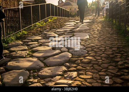 Ein einsamer Fußgänger spaziert in der Nähe des Kolosseums in Rom und wirft einen langen Schatten über große Kopfsteinpflaster einer alten römischen Straße im warmen Sonnenlicht. Stockfoto