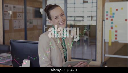 Lächelnde Geschäftsfrau mit Blazer und Tablet im modernen Büro, mit Monitor und Whiteboard Stockfoto