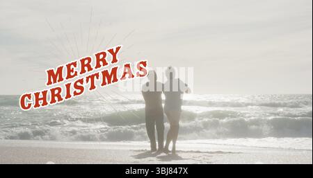 Stehende Seniorenpaare beobachten die Wellen am Sandstrand mit Merry Christmas Overlay Stockfoto