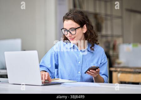 Lächelnde junge Arbeiterin, die ein Mobiltelefon benutzt und im Büro am Schreibtisch sitzt. Stockfoto