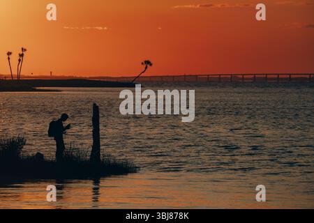 Eine friedliche Sonnenuntergangsszene mit der Silhouette eines Fischers, der seine Schnur in der Nähe von Palmen wirft, mit einer langen Brücke im Hintergrund. Die goldene LIGG Stockfoto
