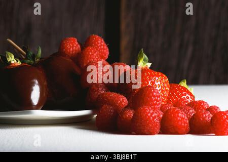 Makroaufnahme von Hochglanz-Erdbeeren aus dunkler Schokolade auf Spießen neben einem Hügel aus saftigen Himbeeren auf einem weißen Teller und rustikaler Kulisse; perfekter Gour Stockfoto